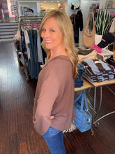 A woman with blonde hair wearing a ribbed, long-sleeve brown top and blue jeans, posing inside a clothing boutique.