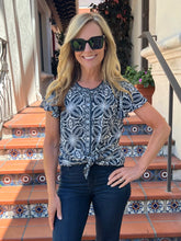 A woman wearing a navy and white patterned short-sleeve top with a front knot detail, paired with dark blue jeans and black sunglasses, standing on tiled outdoor stairs.