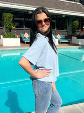 Woman standing by a pool wearing sunglasses and a light blue shirt.