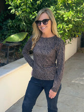 A woman wearing a long-sleeve crewneck knit top with a black and brown abstract pattern, paired with dark jeans and sunglasses, standing outdoors in front of a white wall and green foliage.