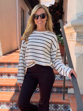A woman wearing a cream and black horizontally striped long-sleeve knit sweater and dark brown pants, standing on tiled outdoor stairs.