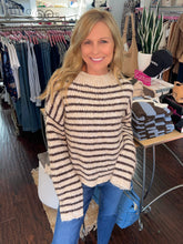 A smiling blonde woman wearing a cream and dark brown horizontally striped knit sweater and blue jeans, standing inside a clothing boutique with racks of garments and a display table in the background.