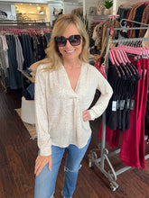 A woman wearing a cream-colored long-sleeve blouse with a subtle floral pattern and blue jeans, standing inside a clothing boutique.