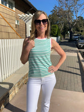 Woman in striped shirt and white pants standing on a sidewalk with a house and trees in the background.