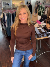 A smiling blonde woman wearing a fitted brown mock neck long-sleeve top and blue jeans, standing inside a clothing boutique.