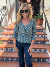A woman wearing a blue and white paisley patterned V-neck top with three-quarter sleeves and dark blue jeans, standing on tiled terracotta steps.