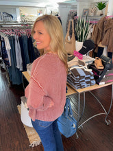 A woman with blonde hair wearing a pink ribbed knit sweater and blue jeans, standing inside a clothing boutique next to a table with folded sweaters and accessories.