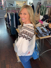A woman smiling and looking back over her shoulder while wearing a cream knit sweater with a brown and beige geometric pattern on the upper chest and sleeves, paired with blue jeans in a clothing boutique.