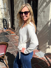 A woman wearing a light beige knit tie-front sweater and blue jeans, standing outdoors on a brick patio in front of a stucco wall.