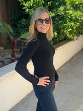 A woman with blonde hair and sunglasses wearing a black ribbed mock neck long-sleeve top and dark blue jeans, posing outdoors on a concrete walkway with green plants and a beige wall in the background.