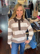 A smiling blonde woman wearing a brown and cream horizontally striped knit polo sweater with blue jeans, standing inside a clothing boutique.