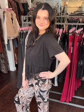 A woman posing in a clothing boutique wearing a black short-sleeve top with a lace hem and black, white, and beige patterned pants, set against a background of clothing racks.
