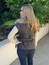 A woman wearing a dark brown diamond-quilted puffer vest with a faux-fur collar, worn over a cream knit sweater and dark blue jeans, standing outdoors against a background of green foliage and a white wall.