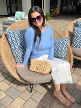 Woman sitting on a wicker chair with blue and gray cushions, holding a woven handbag.