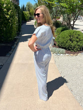 Woman in a light blue outfit standing on a sidewalk with greenery around.