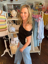 Woman standing wearing a black top off one shoulder in a clothing store with racks of clothes in the background