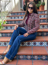 A woman wearing a red and black tweed blazer, blue jeans, and a burgundy top sits on terracotta tiled outdoor stairs with decorative blue and white patterned tiles.