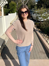 Woman wearing a khaki ribbed knit top standing on a sidewalk with trees and cars in the background.
