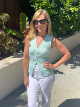 Woman wearing a light green sleeveless top and white pants standing outdoors with plants in the background.