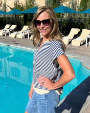 Woman in a striped shirt and sunglasses standing by a pool with lounge chairs and umbrellas in the background.