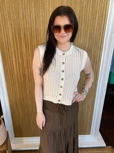 Woman wearing a cream sleeveless top with buttons and a brown skirt, standing against a wooden paneled wall.