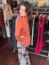 A woman with long dark hair stands in a clothing boutique, looking back over her shoulder. She is wearing a rust-orange long-sleeve mock neck top and black, white, and gray patterned wide-leg pants.