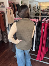 A woman viewed from behind wearing an olive green quilted sleeveless vest over a beige long-sleeve top and blue jeans, standing inside a clothing boutique in front of racks of garments.