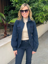 A woman wearing a dark blue denim jacket over a cream ribbed turtleneck sweater and dark blue jeans, standing outdoors in front of green foliage and a beige wall.