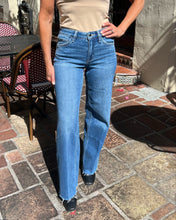 A woman wearing a beige tank top and blue straight-leg jeans stands on a stone patio outdoors. The photo is cropped from the shoulders down, showing the jeans clearly in natural sunlight.
