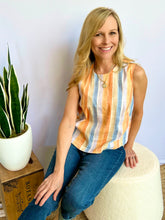 Woman wearing a striped sleeveless top and jeans standing in a room with a plant and ottoman.