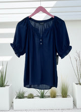 Navy blue blouse with button details on a hanger against a white background with plants.