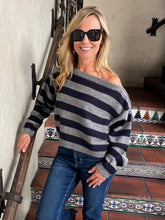 A woman wearing a gray and navy blue horizontally striped off-the-shoulder sweater, blue jeans, and black sunglasses, posing on a tiled staircase.