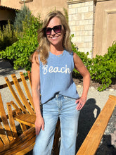 Woman wearing a blue 'Beach' tank top standing outdoors near wooden chairs.