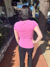 Person wearing a lilac shirt and black pants standing on a sidewalk with trees and buildings in the background.
