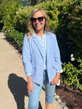 Woman wearing a light blue blazer and jeans standing outdoors with greenery in the background