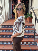 A woman wearing a brown and white horizontally striped knit top and brown pants, standing on tiled outdoor stairs.