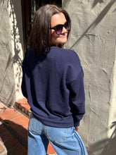 A woman wearing a navy blue crewneck sweatshirt and light blue jeans, photographed from behind in an outdoor setting against a textured stucco wall.