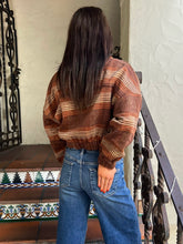 A woman viewed from behind wearing a brown and beige plaid cropped sweater and blue jeans, standing on an outdoor staircase with a tiled step and black metal railing.
