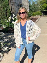 Woman wearing sunglasses, a white cardigan, blue top, and jeans standing outdoors.