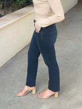 A person wearing dark navy blue straight-leg jeans, a beige ribbed long-sleeve top, and beige block-heel sandals, standing on a concrete patio.