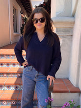 A woman wearing a dark navy ribbed knit V-neck sweater and blue jeans stands on outdoor terracotta steps in front of a white stucco building.