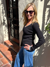 A woman wearing a black ribbed long-sleeve sweater with white trim on the V-neck and cuffs, paired with blue jeans, standing outdoors against a stucco wall.