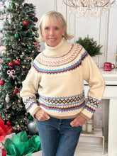 Woman wearing a cream turtle neck sweater with a colorful pattern in a festive setting with a decorated Christmas tree.