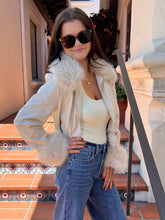 A woman wearing a cream faux-fur trimmed jacket over a cream bodysuit and blue jeans, standing on outdoor tiled steps.