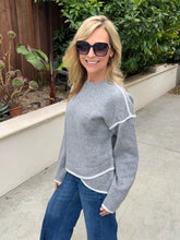 A woman wearing a light gray knit sweater with white contrast trim and blue jeans, standing outdoors on a concrete walkway with green plants and a wooden fence in the background.
