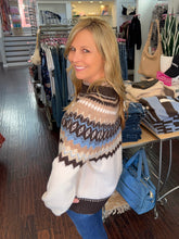 A woman with blonde hair smiling and posing inside a clothing boutique, wearing a white knit sweater with a brown, beige, and light blue geometric pattern.