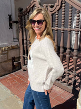 A woman wearing a light beige knit sweater and blue jeans stands outdoors in front of a decorative brown metal railing and tiled ground.
