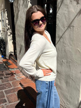 A woman wearing a cream-colored long-sleeve knit sweater, blue jeans, and dark sunglasses, posing outdoors against a textured stucco wall.