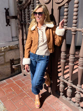 A woman wearing a tan suede moto jacket with white faux-fur trim, a cream henley top, blue jeans, and tan heeled sandals, posing outdoors next to a decorative metal gate.
