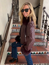 A woman wearing a brown leather bomber jacket with dark fur trim, blue jeans, and black sunglasses, posing on a tiled staircase.